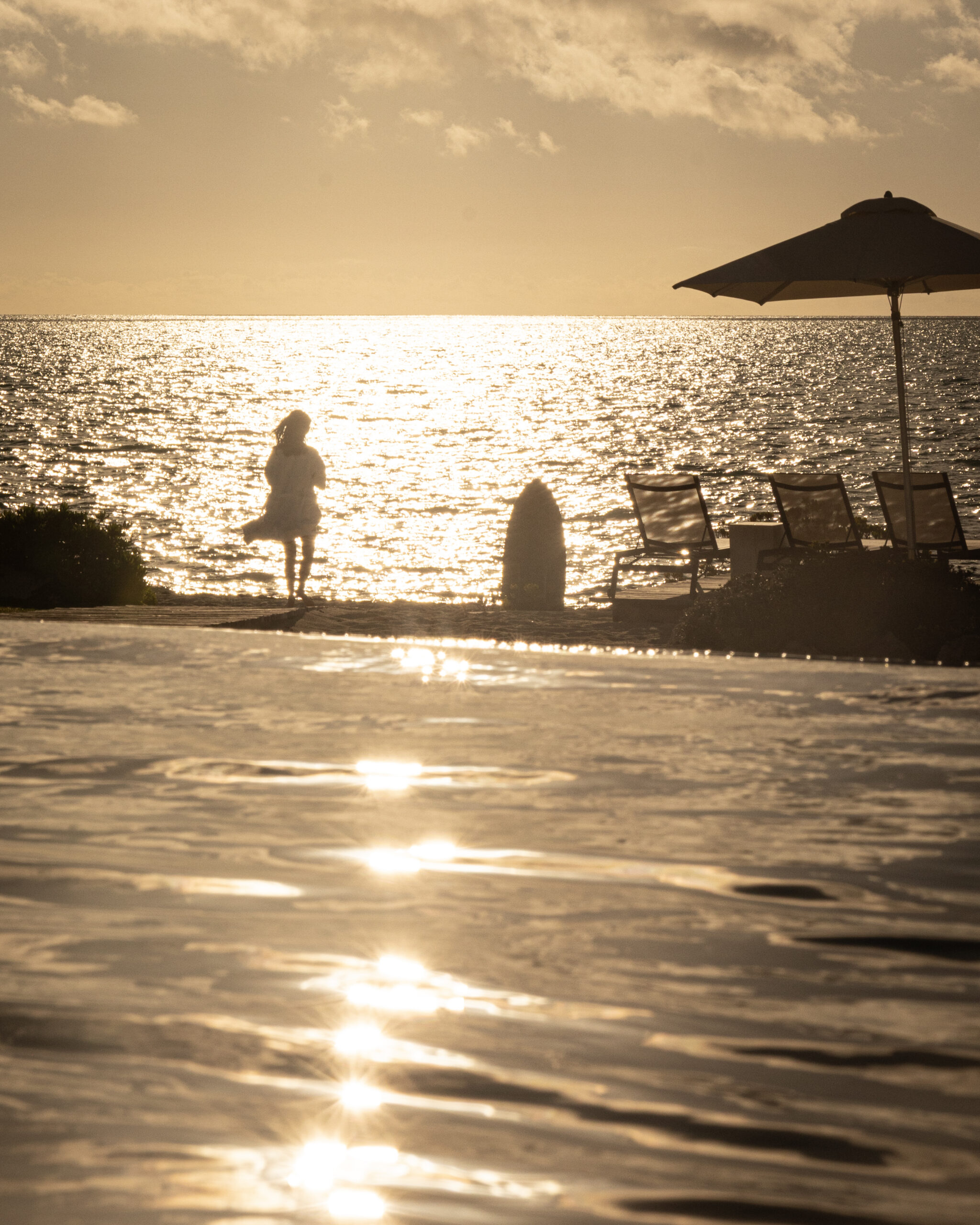 Silhouette of a person standing at the edge of an infinity pool overlooking the ocean at sunset, with lounge chairs and a beach umbrella nearby.