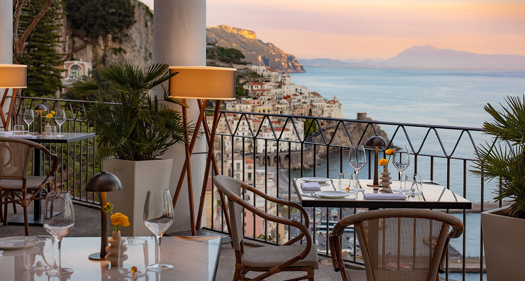Elegant cliffside restaurant terrace with set dining tables, wrought iron railing, and sweeping views of a coastal town and sea at sunset.