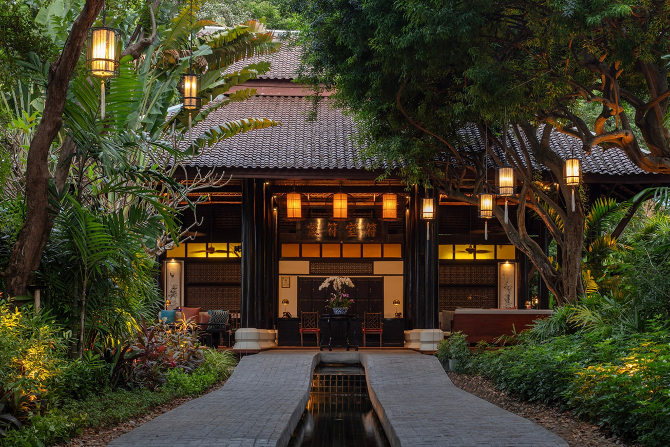 Warmly lit traditional resort entrance pavilion at dusk, with a reflecting pool pathway, glowing lanterns, and lush tropical garden surroundings.