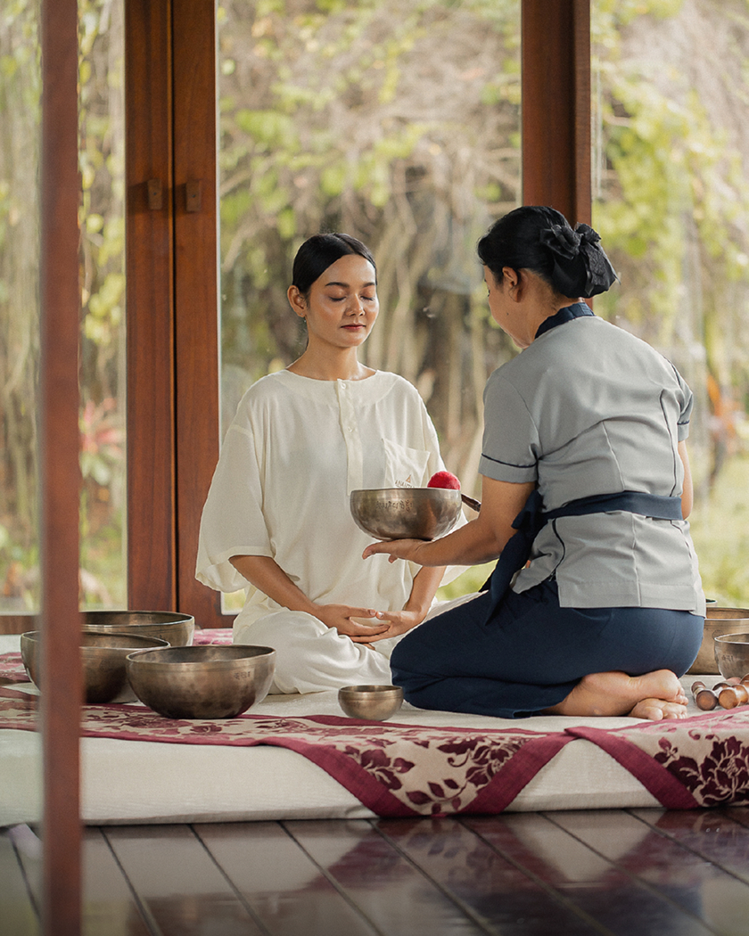 Spa therapist presenting a singing bowl to a relaxed guest during a wellness ritual in an open-air treatment pavilion surrounded by nature.