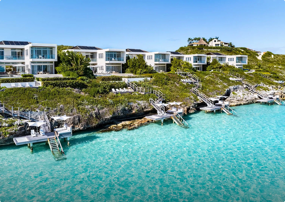 Aerial view of modern white luxury villas perched on a rocky cliffside overlooking crystal-clear turquoise water, with private docks and staircases leading down to the sea.