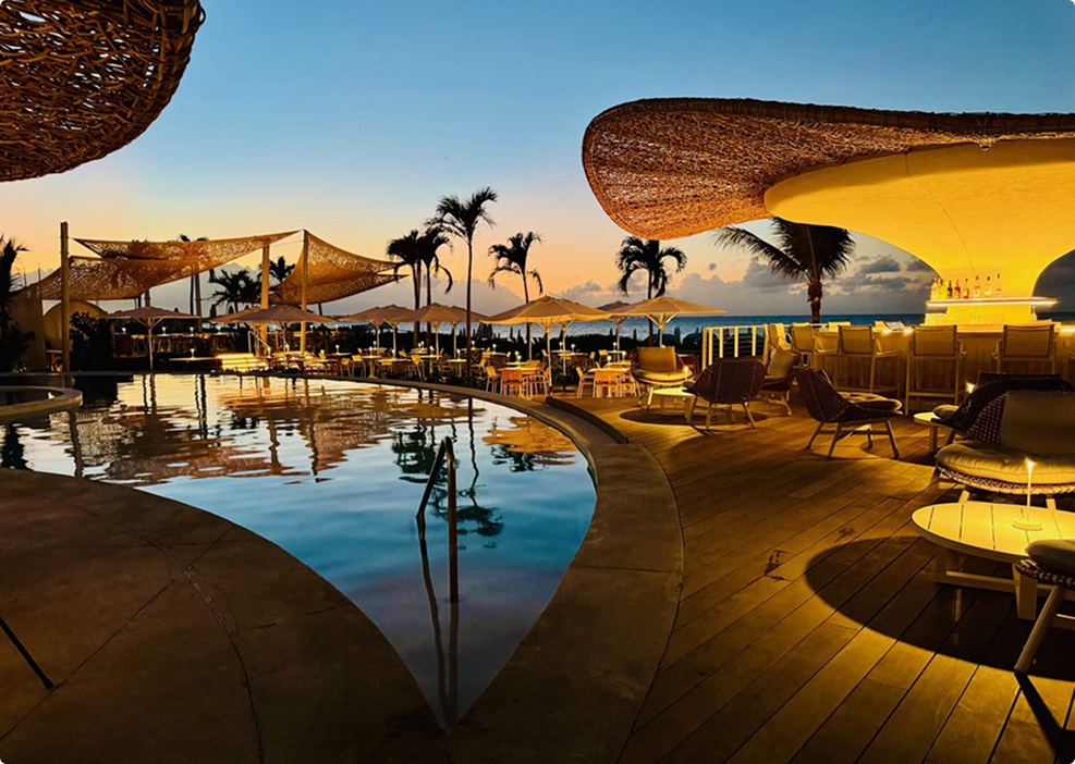 Upscale beachfront pool bar at sunset with woven rattan shade structures, glowing yellow lighting, lounge seating, and palm tree silhouettes reflecting in the water.