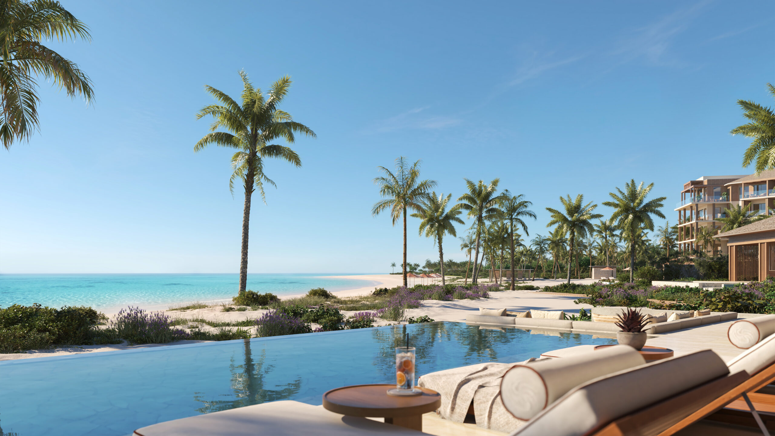 A luxury resort infinity pool with cream loungers in the foreground, reflecting tall palm trees and a white sand beach with turquoise water under a clear blue sky.