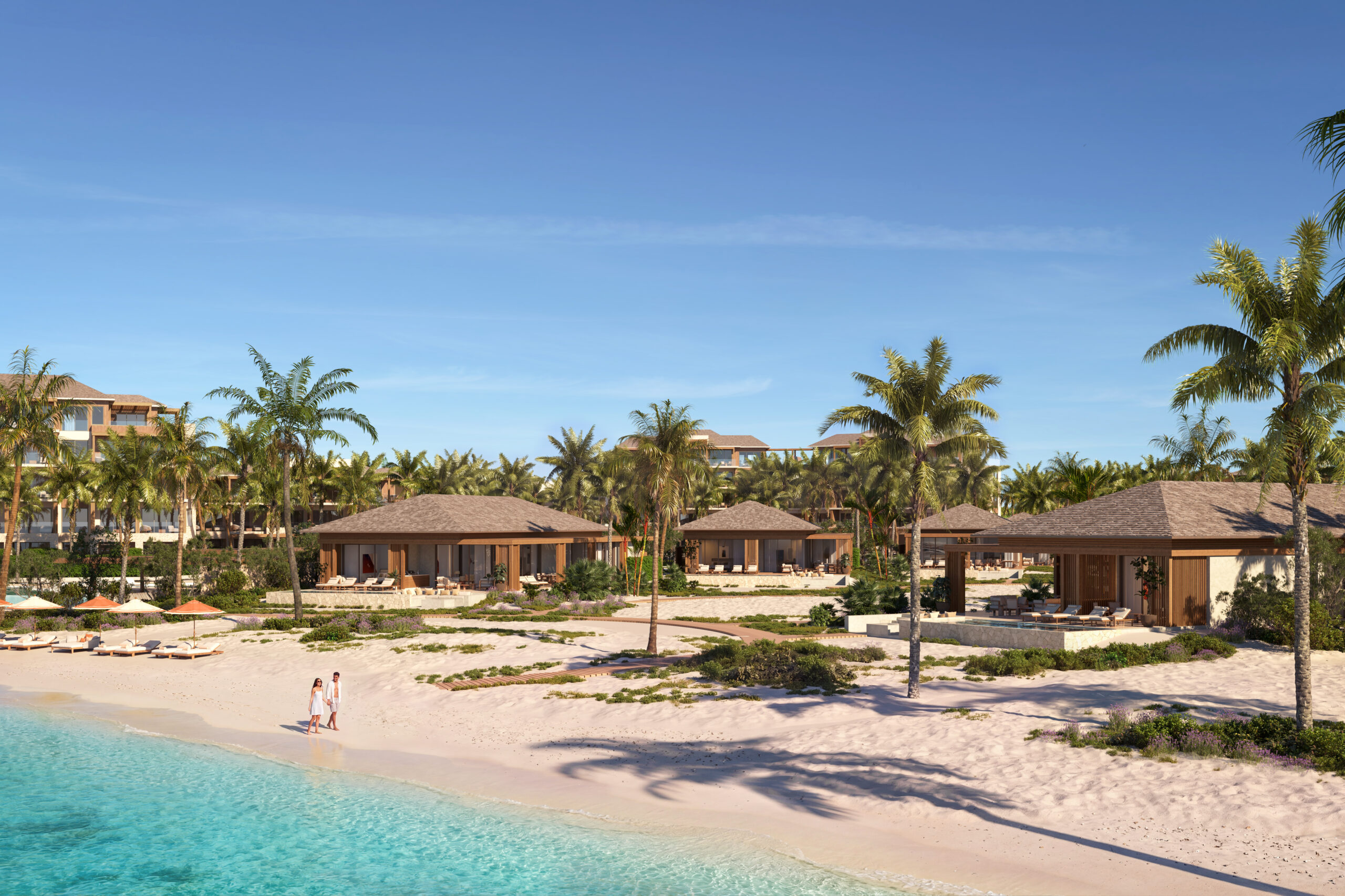 view from the sea looking onto th full Anantara turks and caicos property. Low buildings and lush greenery give the sense that this place has been untouched. A couple walks leisurely down the beach.